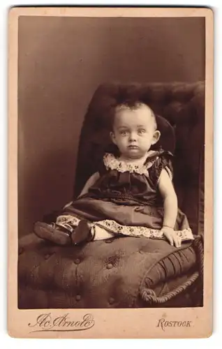 Fotografie Ad. Arnold, Rostock, Portrait niedliches Kleinkind im hübschen Kleid auf Sessel sitzend