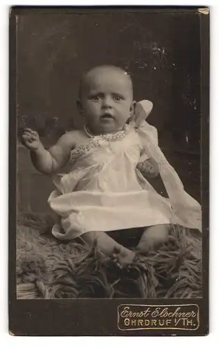 Fotografie Ernst Elschner, Ohrdruf i / Th., Portrait niedliches Baby im weissen Kleid auf Fell sitzend