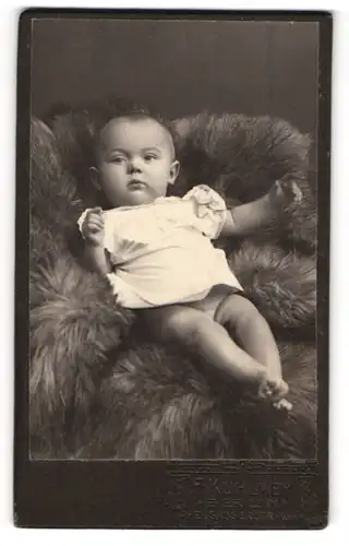Fotografie Franz Kuhlmey, Berlin-N, Portrait niedliches Baby im weissen Hemd auf Fell liegend