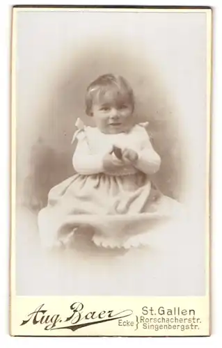 Fotografie Aug. Baer, St. Gallen, Portrait niedliches Kleinkind im hübschen Kleid auf Fell sitzend