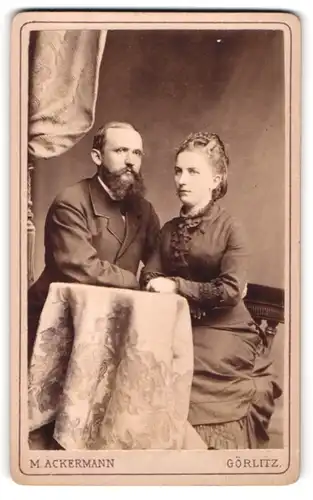 Fotografie M. Ackermann, Görlitz, Portrait bürgerliches Paar in hübscher Kleidung am Tisch