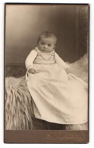 Fotografie Max Petermann, Leipzig-Kl. Zschocher, Portrait niedliches Baby im langen Kleid auf Fell sitzend