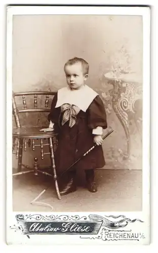 Fotografie Atelier Gliese, Reichenau i / S., Portrait hübsch gekleidetes Kind mit Peitsche an Stuhl gelehnt