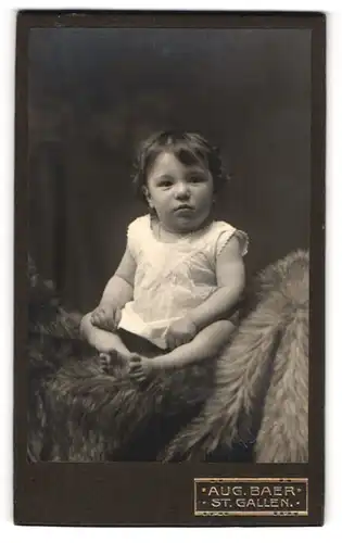 Fotografie Aug. Baer, St. Gallen, Portrait niedliches Kleinkind im weissen Hemd auf Fell sitzend