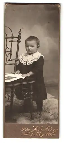 Fotografie Friedr. Kolby, Plauen i / V., Portrait niedliches Kleinkind im hübschen Kleid mit Foto an Stuhl gelehnt