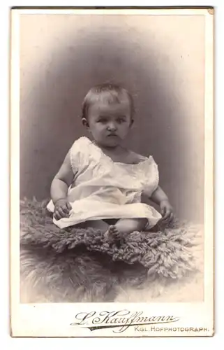 Fotografie L. Kauffmann, Bad Kreuznach, Portrait niedliches Kleinkind im weissen Hemd auf Fell sitzend