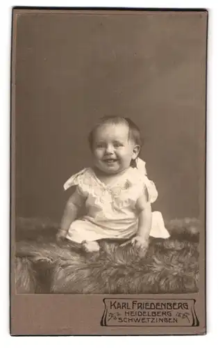 Fotografie Karl Friedenberg, Heidelberg, Portrait niedliches Kleinkind im weissen Hemd auf Fell sitzend