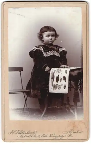 Fotografie H. Haberlandt, Berlin, Portrait kleines Mädchen mit Buch am Tisch stehend