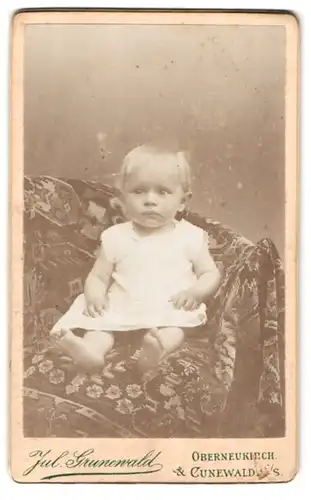 Fotografie Julius Grunewald, Cunewalde / Sachsen, Portrait niedliches Kleinkind im weissen Hemd auf Decke sitzend