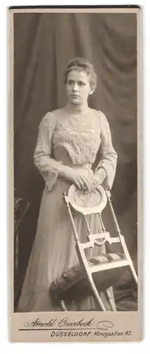 Fotografie Arnold Overbeck, Düsseldorf, Königsallee 43, Frau im verzierten Kleid steht an einem Stuhl