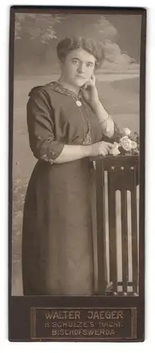 Fotografie Walter Jaeger, Bischofswerda, Frau mir Schmuck verziert steht im Kleid und Blumen an einem Stuhl