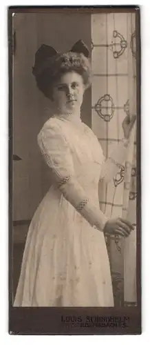 Fotografie Louis Schindhelm, Ebersbach i. S., Frau im weissen Kleid mit schwarzer Schleife im Haar steht am Fenster
