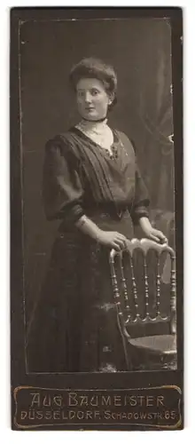Fotografie Aug. Baumeister, Düsseldorf, Schadowstrasse 65, Frau im schwarzen Kleid mit weisser Bluse steht an einem Stuhl