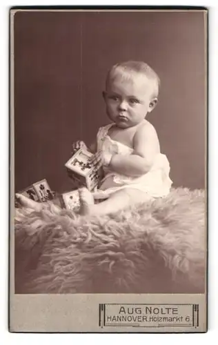 Fotografie Aug. Nolte, Hannover, Holzmarkt 6, Portrait niedliches Baby im weissen Hemd mit Würfeln auf Fell sitzend