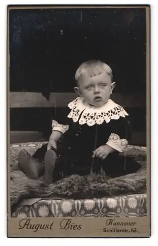 Fotografie August Bies, Hannover, Schillerstrasse 52, Portrait niedliches Kleinkind in hübscher Kleidung auf Fell sitzen