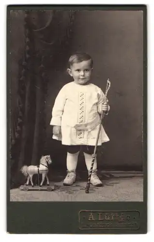 Fotografie A. Lüttge, L.-Leutzsch, Hauptstrasse 34, Junge im weissen Gewand mit einem Spielpferd