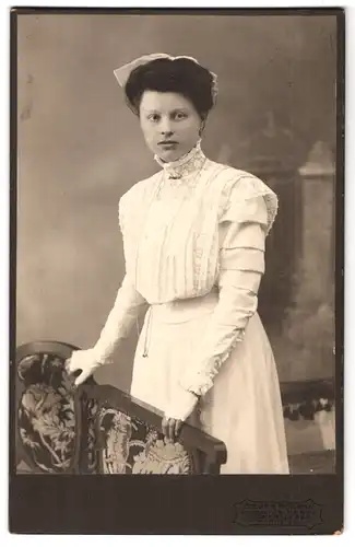 Fotografie Atelier Modern, Aschersleben, Portrait von M. Gieseler 1909 im weissen Kleid mit Haube an einem Stuhl