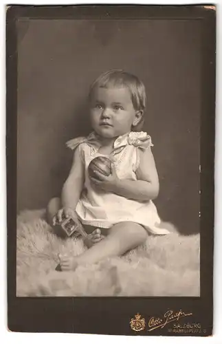 Fotografie Otto Paap, Salzburg, Mirabellplatz 6, Portrait niedliches Kleinkind im weissen Hemd mit Ball auf Fell sitzend