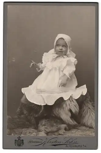 Fotografie Kersten Sohn, Altenburg S / A., Portrait nieldiches Baby im weissen Kleid auf Fell sitzend