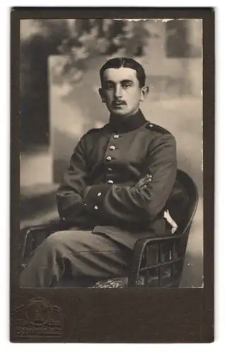 Fotografie Hermann Tietz, München, Bahnhofplatz, Portrait bayrischer Soldat in Uniform auf Rattan Stuhl