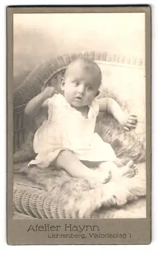 Fotografie Atelier Haynn, Berlin-Lichtenberg, Viktoriaplatz 1, Portrait neidliches Kleinkind im Hemd auf Fell sitzend