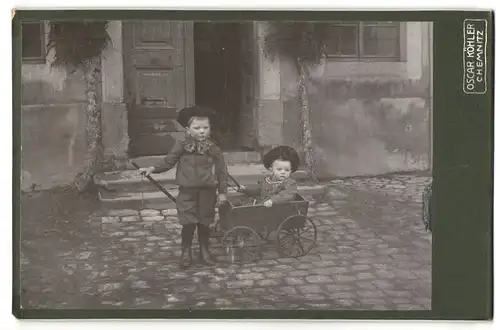 Fotografie Oskar Köhler, Chemnitz, Portrait zwei niedliche Kleinkinder mit Bollerwagen vor dem Hauseingang