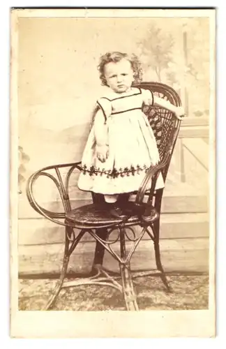 Fotografie unbekannter Fotograf und Ort, niedliches kleines Mädchen im hellen Kleid auf einem Stuhl stehend