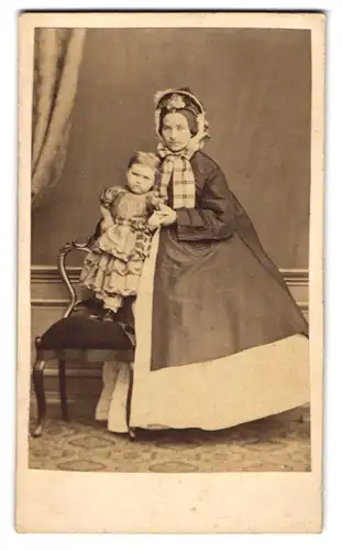 Fotografie E. G. Murken, Varel, Mutter im Winterkleid mit Haube und Tochter auf dem Stuhl