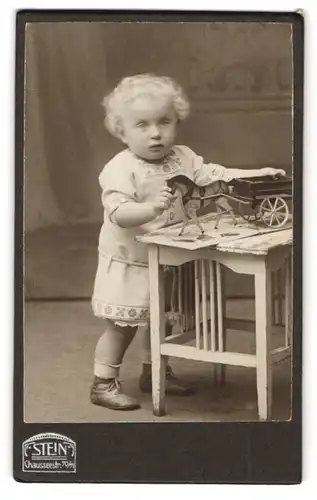 Fotografie F. Stein, Berlin, niedliches blondes Kind mit Holzpferd und Karren auf dem Tisch