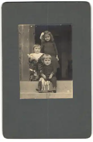 Fotografie unbekannter Fotograf und Ort, drei niedliche Kinder mit Spielzeug Pferd und Puppe im Arm