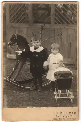 Fotografie Th. Dittmar, Herzberg a. E., zwei niedliche Kinder mit Puppenwagen und Schaukelpferd im Garten