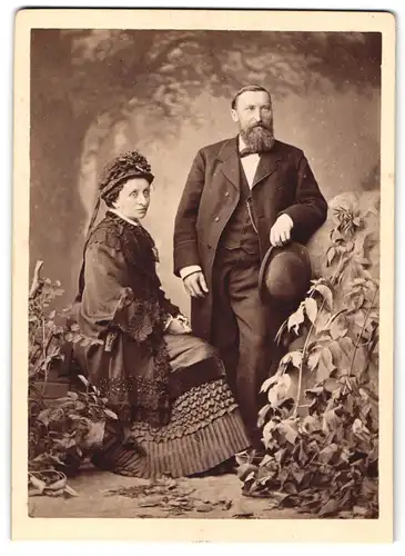 Fotografie unbekannter Fotograf und Ort, älteres Paar im Gründerzeit Kleid und Anzug mit Melone, Studiokulisse