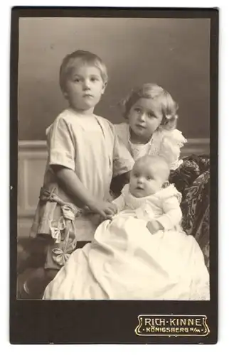 Fotografie Rich Kinne, Königsberg n. M., Niedliche Geschwisterkinder in weisser Kleidung mit aufgeregtem Baby