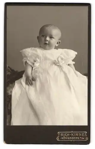 Fotografie Rich. Kinne, Königsberg n. M., Süsses Baby im weissen Kleid schaut mit grossen Augen erstaunt zur Seite