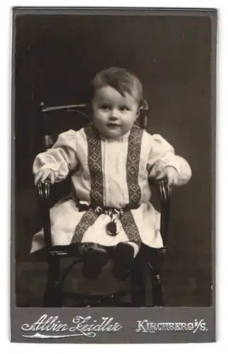 Fotografie Albin Zeidler, Kirchberg, Neumarkt 238, Süsses Kleinkind im weissen Kleidchen sitzt erfreut auf einem Stuhl