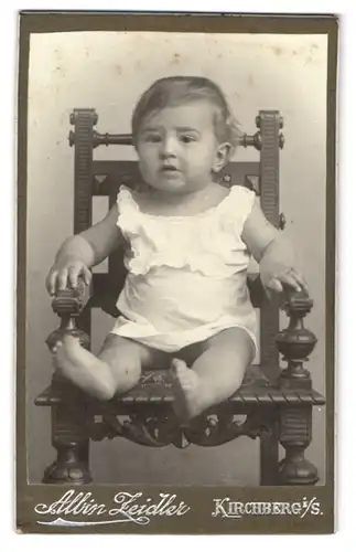 Fotografie Albin Zeidler, Kirchberg, Neumarkt 238, Niedliches Baby im weissen Kleid sitzt unglücklich auf einem Stuhl