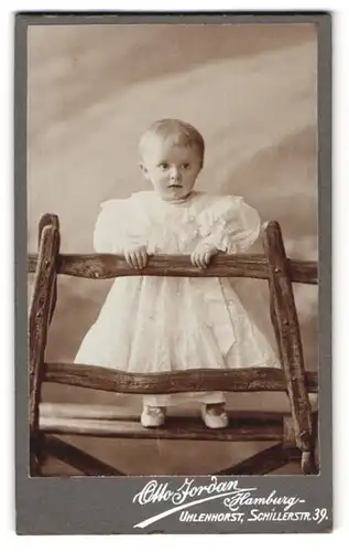 Fotografie Otto Jordan, Hamburg, Schillerstr. 39, Niedliches Kleinkind im weissen Kleid steht auf einer Bank