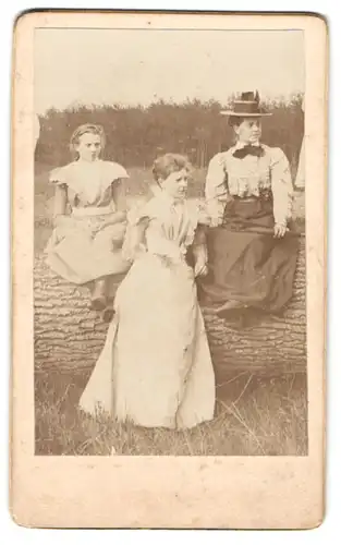 Fotografie unbekannter Fotograf und Ort, drei junge Damen in Sommerkleidern mit Hut und Schirm auf grossem Baumstamm