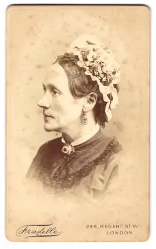 Fotografie Fradelle, London, ältere Dame im dunklen Kleid mit Brosche, Ohringen und Blumenhut