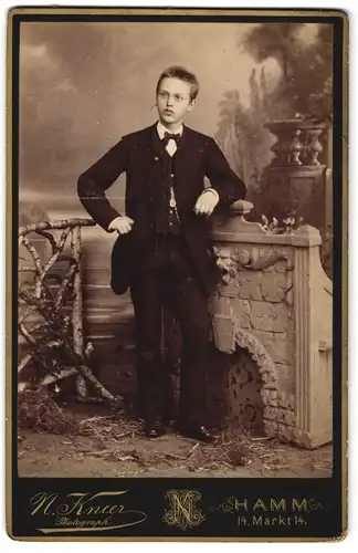 Fotografie N. Kneer, Hamm a. d. Lippe, junger Knabe im Anzug mit Fliege vor Gründerzeit Kamin