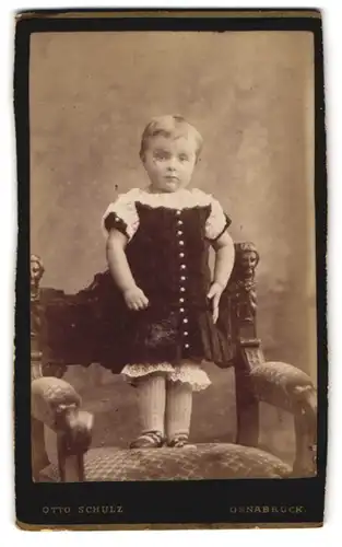 Fotografie Otto Schulz, Osnabrück, Hasestr. 59, Niedliches Kind im schwarzen Kleid steht auf einem Stuhl