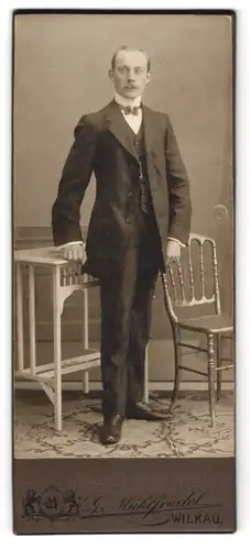 Fotografie Gustav Mühlfriedel, Wilkau i. S., Feiner Herr im Anzug steht zwischen Tisch und Stuhl