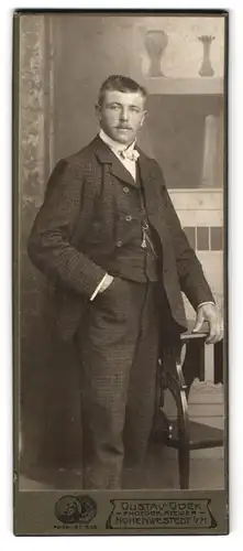 Fotografie Gustav Gock, Hohenwestedt i. H., Junger Herr mit kariertem Anzug steht an Tisch