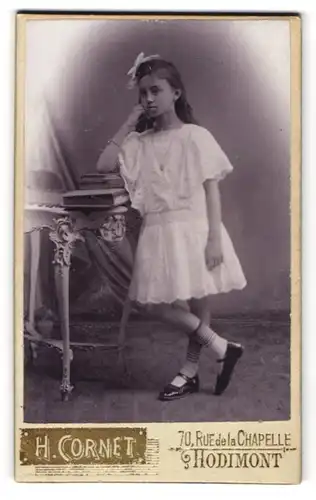 Fotografie H. Cornet, Hodimont, 70 Rue de la Chapelle, Junges Mädel im weissen Kleid mit Schleife gelangweilt nebst Tisch