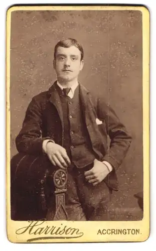 Fotografie Harrison, Accrington, Junger Herr im dunklen Anzug mit Krawatte und Anstecktuch nebst Stuhl