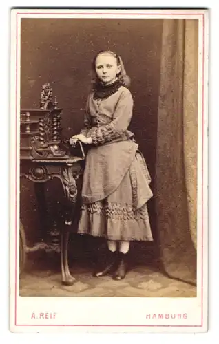 Fotografie A. Reif, Hamburg, Bazar 16, Junges Mädel im hellen Kleid mit schwarzer Spitze und Haarreif nebst Tisch