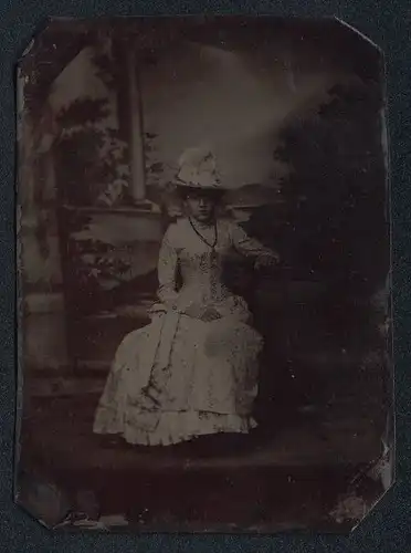 Fotografie Ferrotypie junge Frau im weissen Kleid mit Blumenmuster und Hut vor einer Studiokulisse