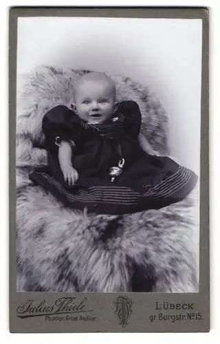 Fotografie Julius Thiele, Lübeck, gr. Burgstr. 15, Lächelndes Kleinkind in zu grossem Kleid auf Fell