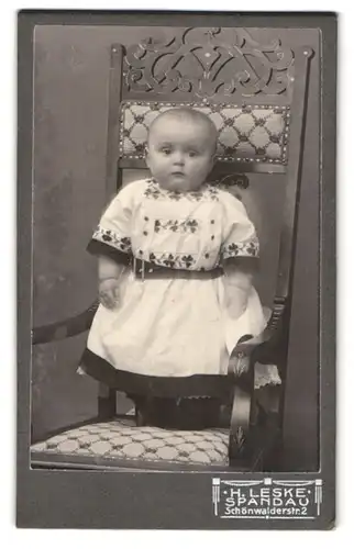 Fotografie H. Leske, Spandau, Schönwalderstr. 2, Baby in einem trachtenähnlichen Kleid auf einem Stuhl stehend