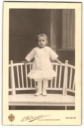 Fotografie S. Weitzmann, Wien, niedliches kleines Mädchen im weissen Kleid posiert stehend auf einer Bank